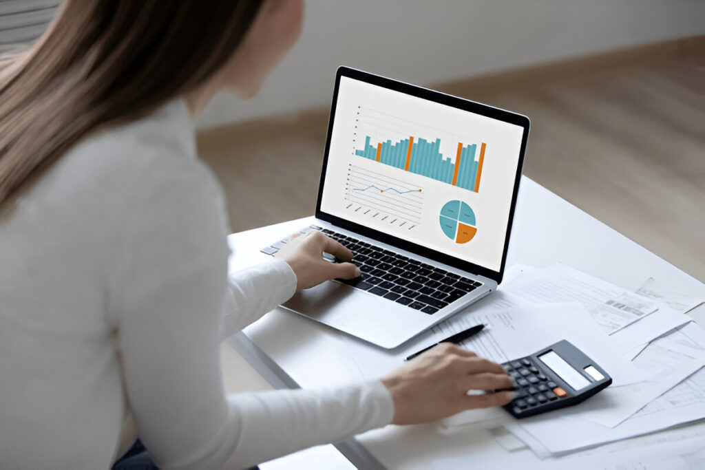 A person in a white shirt works on a laptop displaying charts and graphs, while using a calculator. Papers are scattered on the white desk, hinting at an environment focused on data analysis or financial tasks related to budgeting for cheap travel for students.