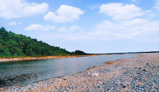 A serene river flows alongside a pebbly shore under a bright blue sky with scattered clouds. A green, forested hill rises on the left, while the opposite bank stretches into the distance, creating a peaceful natural scene.