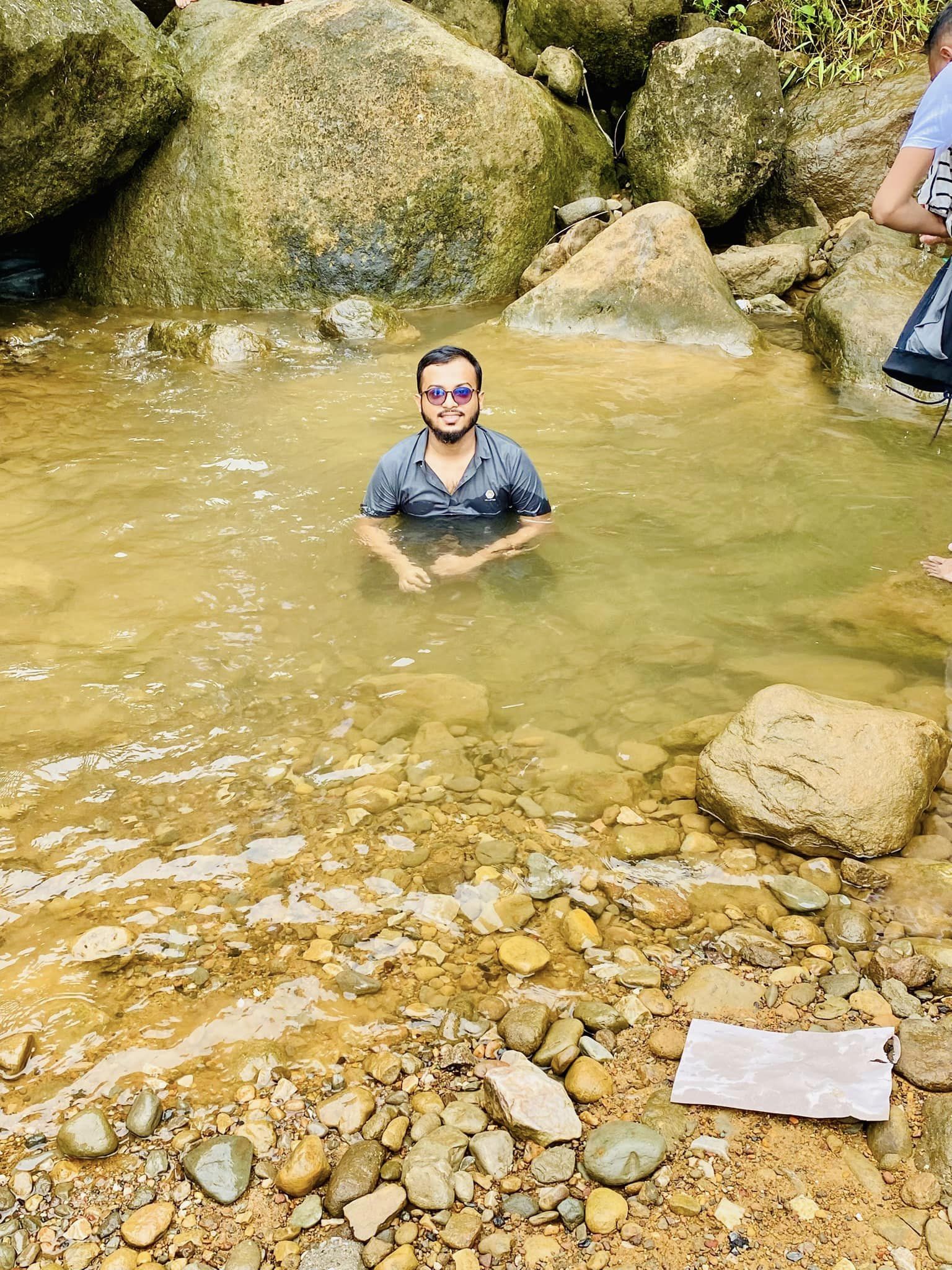 A person wearing sunglasses smiles while sitting in a shallow, rocky stream surrounded by large boulders. The water is clear and the surrounding area is natural and serene. A piece of white paper is on a rock nearby.