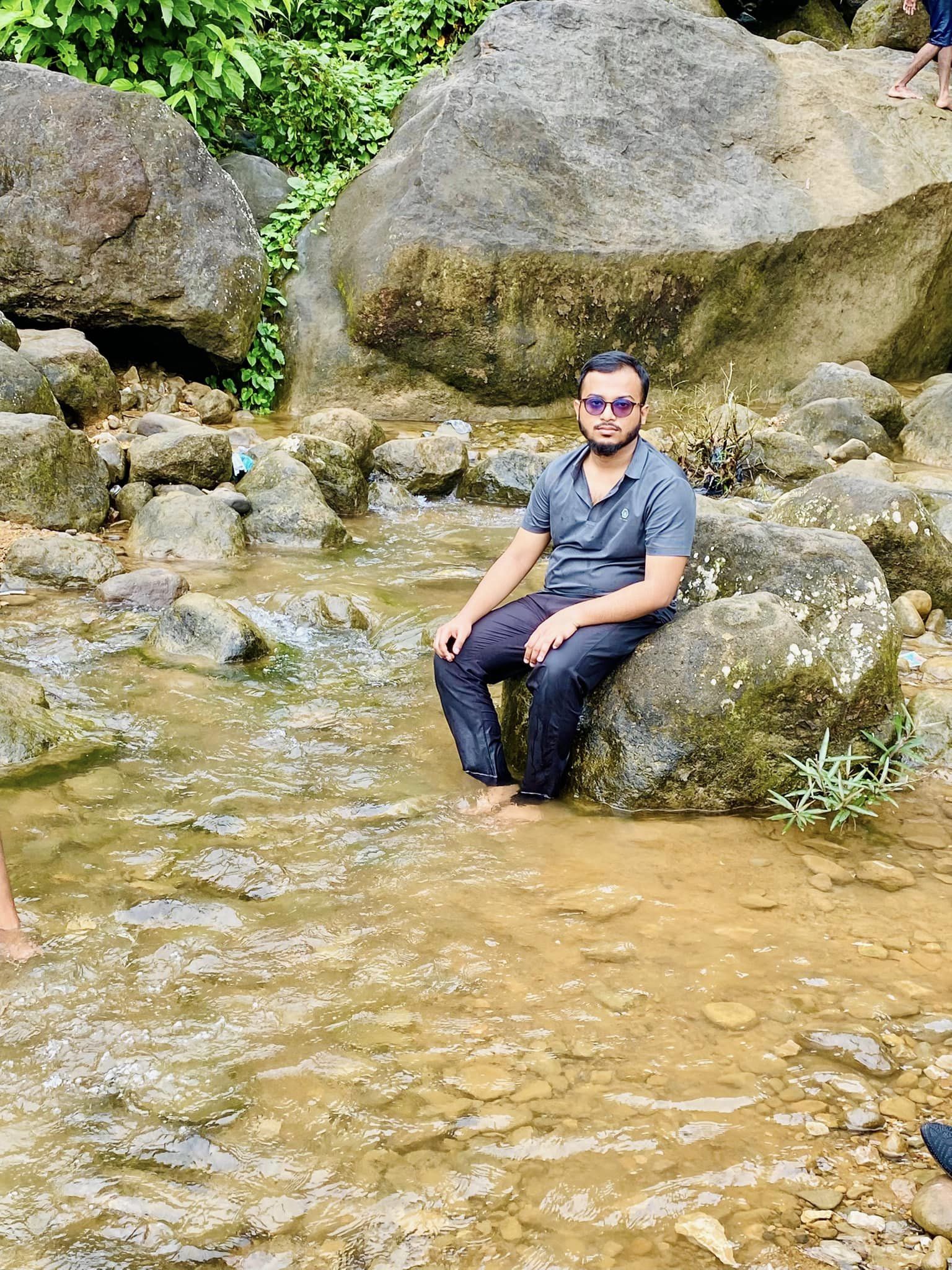 A person wearing sunglasses and casual clothing sits on a rock in a shallow stream surrounded by large rocks and greenery. The water is clear, revealing the rocky streambed.