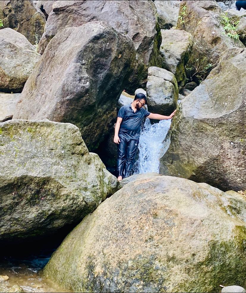 A person wearing a cap and wet clothes leans against large rocks, standing in a stream with water flowing around them. The natural setting is surrounded by moss-covered boulders and greenery.