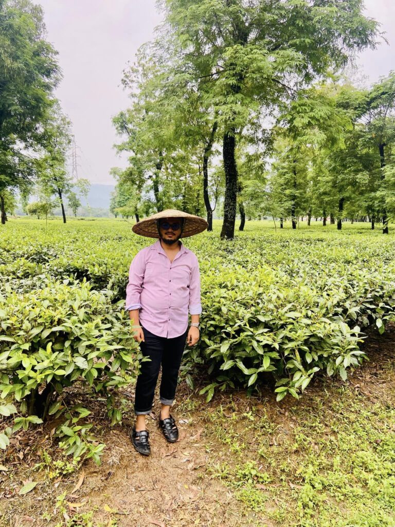 A person wearing a large straw hat and sunglasses stands in a lush tea garden—an ideal destination for travel-loving students. Dressed in a pink shirt and black pants, they are surrounded by vibrant green tea plants and tall trees, capturing the essence of nature's classroom.