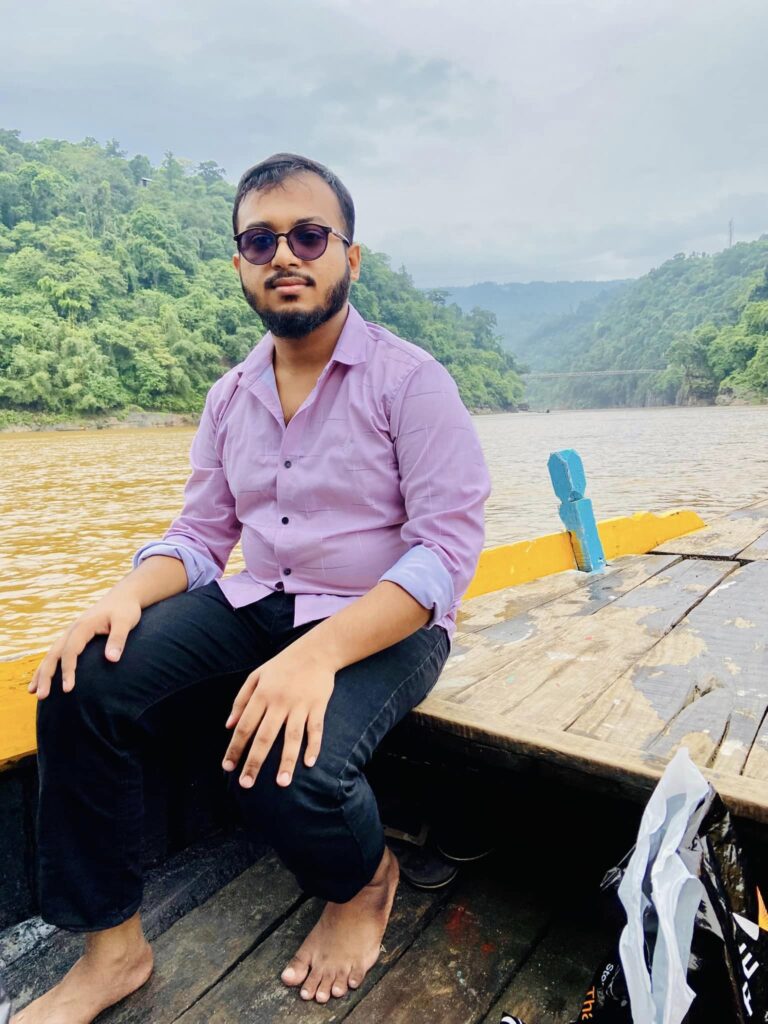 A person wearing a purple shirt and black pants sits barefoot on a boat with a river and lush green hills in the background under a cloudy sky.