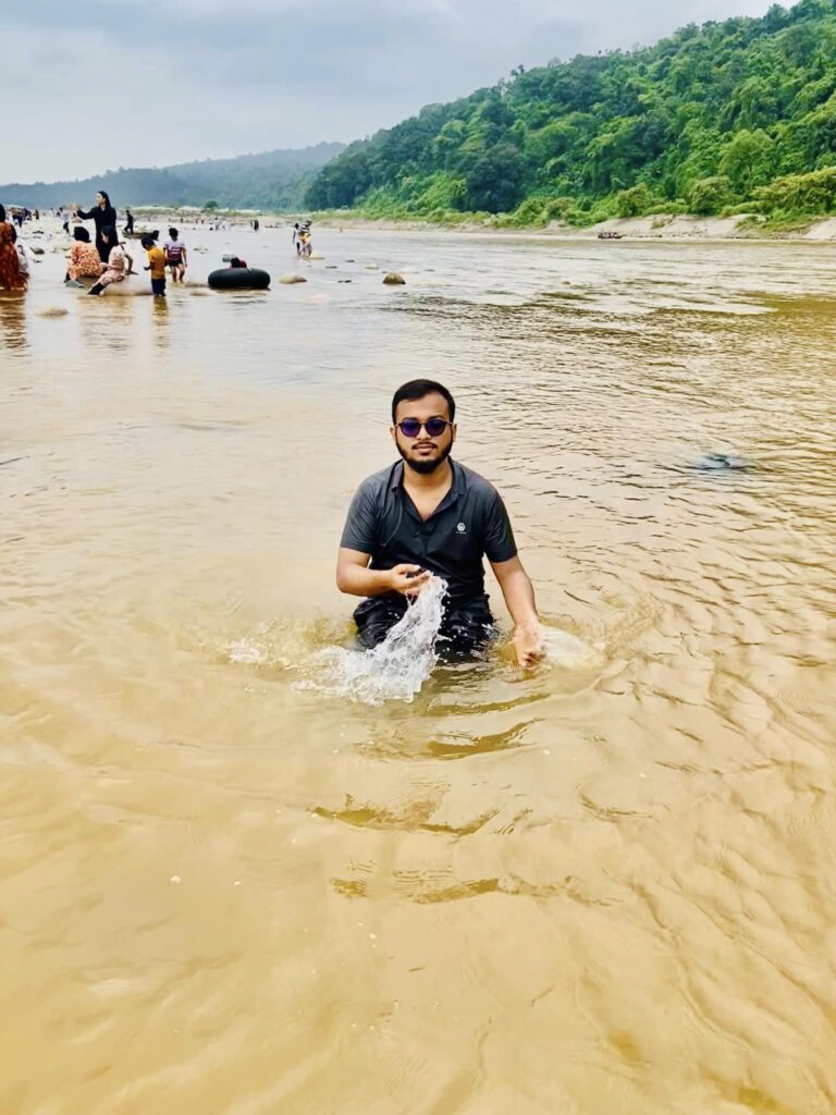 A person wearing sunglasses and a dark shirt kneels in shallow, sandy water, splashing with one hand. In the background, people are gathered by the waters edge, and lush green hills are visible under a cloudy sky.