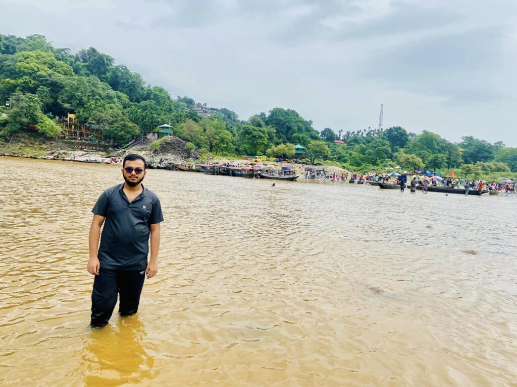 A man stands in shallow, muddy water with distant hills and lush green trees as a backdrop, an intriguing setting for travel for students. Crowds gather near a riverside structure under a cloudy sky, adding to the atmosphere of exploration and discovery.