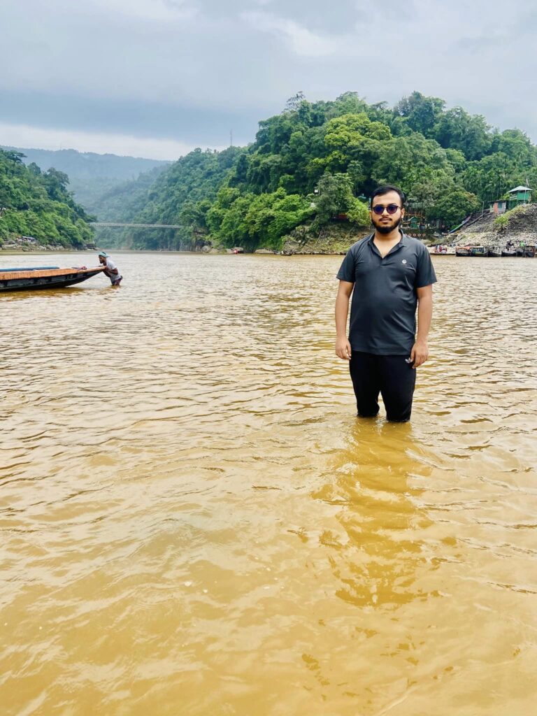 A man stands in a river with knee-deep water, wearing sunglasses and a dark shirt. In the background, green hills rise under a cloudy sky, and a wooden boat floats nearby.