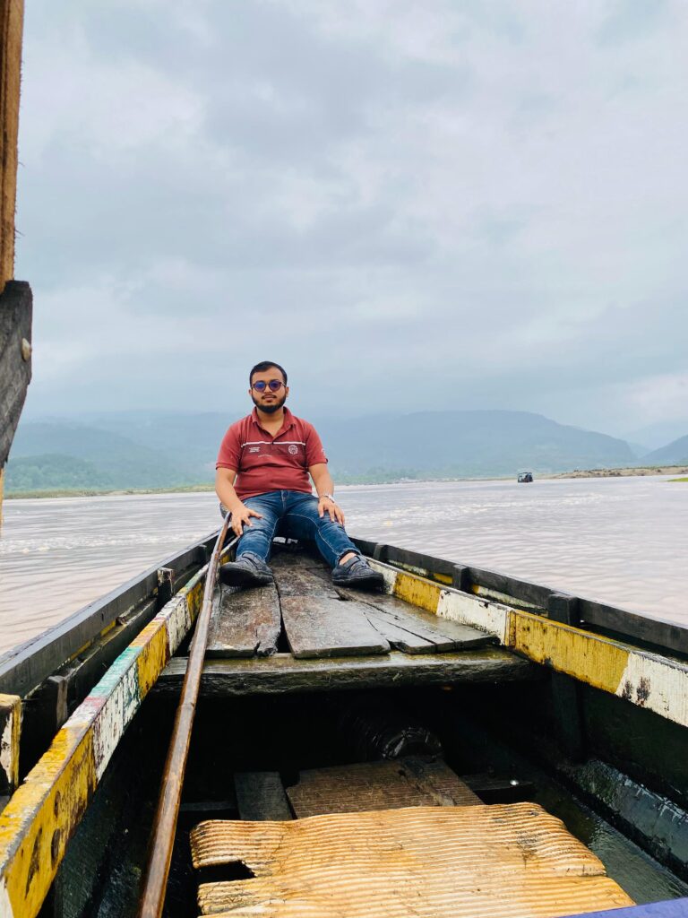 A man wearing a red shirt and sunglasses sits on the deck of a wooden boat, holding a pole. The boat is on a calm body of water with a backdrop of misty mountains and cloudy skies.