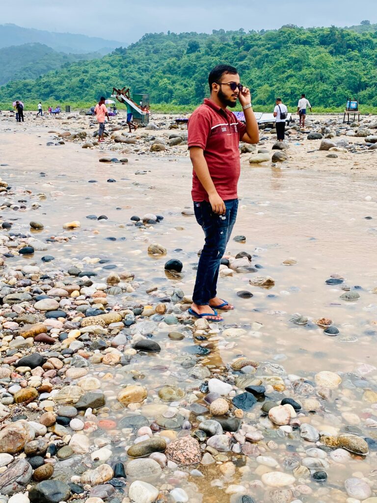 A man in a red shirt and jeans stands on a rocky riverbank, talking on a phone. People in the background walk along the river, and green hills are visible under a cloudy sky.