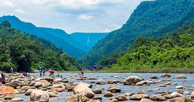 A scenic river winds through a lush, green valley embraced by mountains, perfect for travel-savvy students. Explorers wander the rocky riverbank under a partly cloudy sky, while verdant foliage blankets the land, creating a serene and inspiring natural classroom.