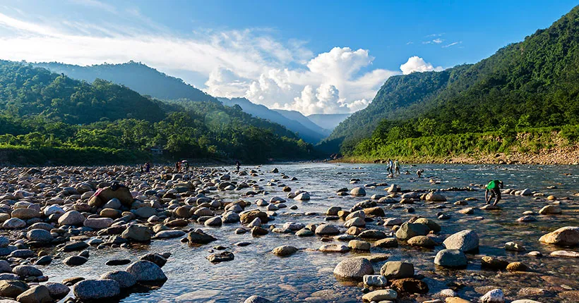 A serene river flows through a rocky bed surrounded by lush green mountains, perfect for travel for students. People are scattered along the river, some standing in the water. The sky is blue with fluffy white clouds, adding to the natural beauty of this ideal student getaway.