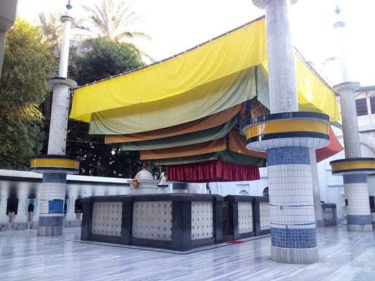 A tomb covered with a colorful canopy in a courtyard. The structure features tiled columns and a marble floor. Surrounding trees and plants provide shade, and the open setting suggests a peaceful, possibly religious site.