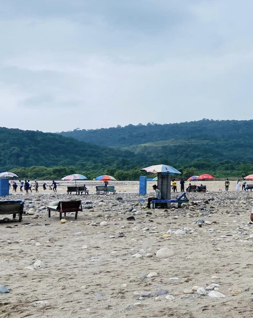 A sandy beach with scattered rocks and a few colorful umbrellas. People are walking in the distance. There are a couple of small structures and a green mountain range in the background under a cloudy sky.