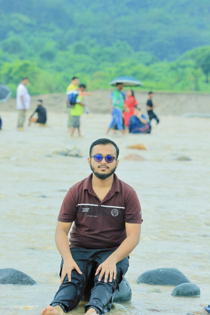 A person wearing sunglasses and a dark shirt sits on rocks by a shallow river. In the background, people walk and stand with umbrellas on a sandy area, with lush green hills visible.