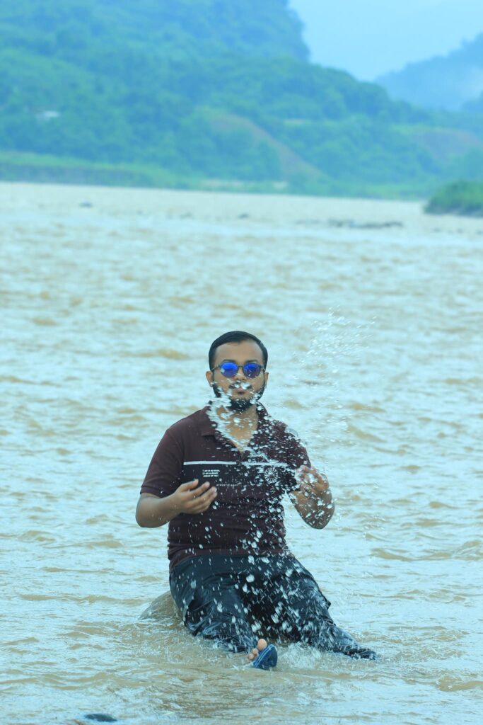 A person wearing sunglasses sits in a river, splashing water with one hand and holding a phone in the other. The background features lush green hills under a cloudy sky.